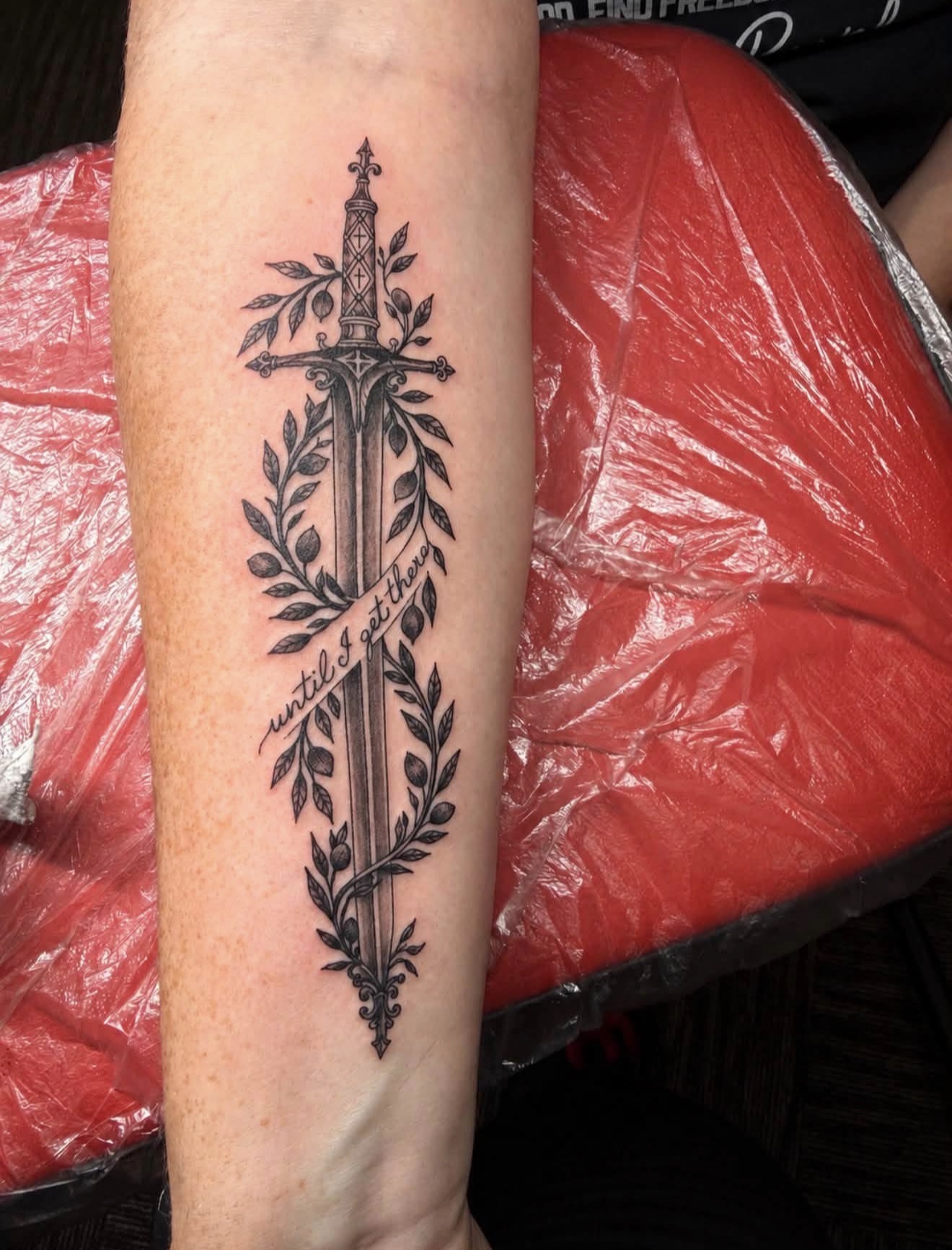 Sword framed by leafy branches and script on a forearm, tattooed by Joseph Rojas